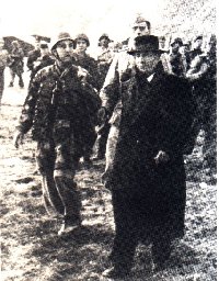 Mussolini indossa un ampio soprabito nero sopra un abito blu scuro troppo largo, ed ha un cappello calato sugli occhi.